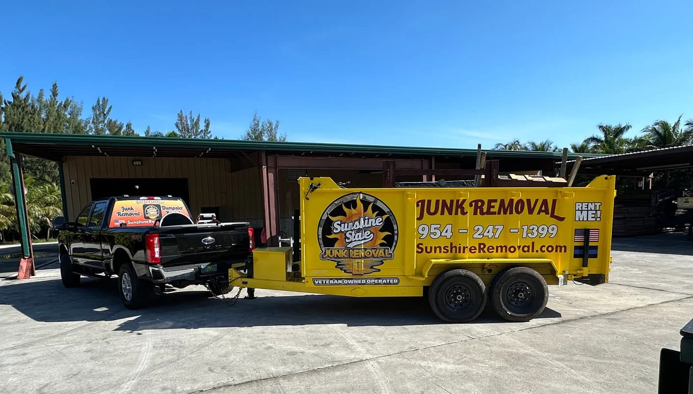 Black pickup truck hitched to a yellow Sunshine State Junk Removal dump trailer under a metal carport